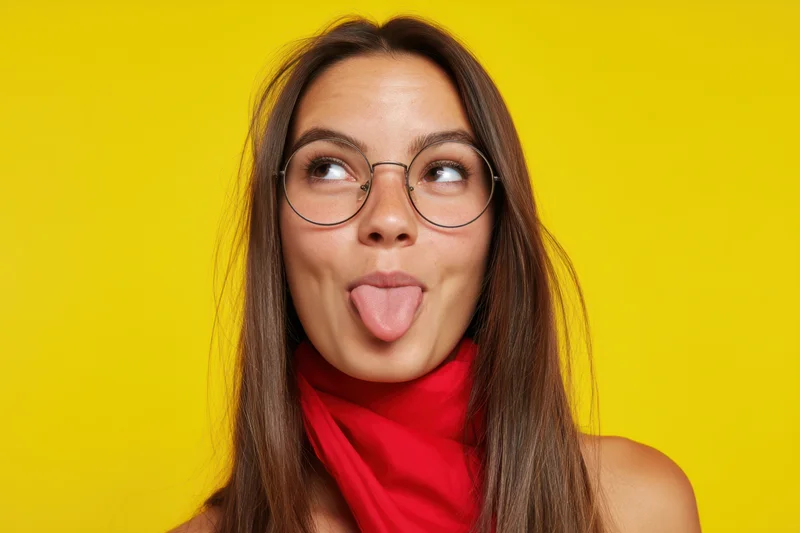 Young woman with long brown hair sticking out her tongue while wearing round glasses and a red scarf against a yellow background