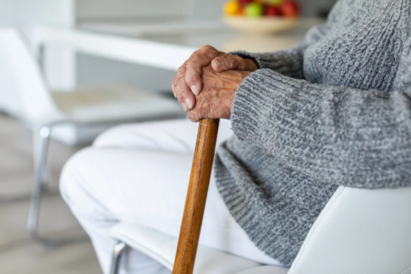 Elderly woman gripping wooden cane while seated in white chair at home
