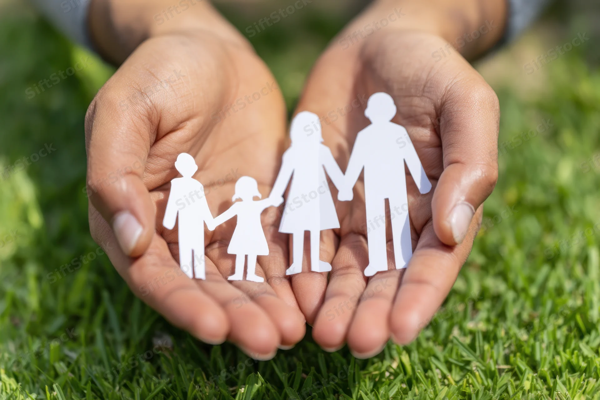Hands holding paper cutout family figures over green grass