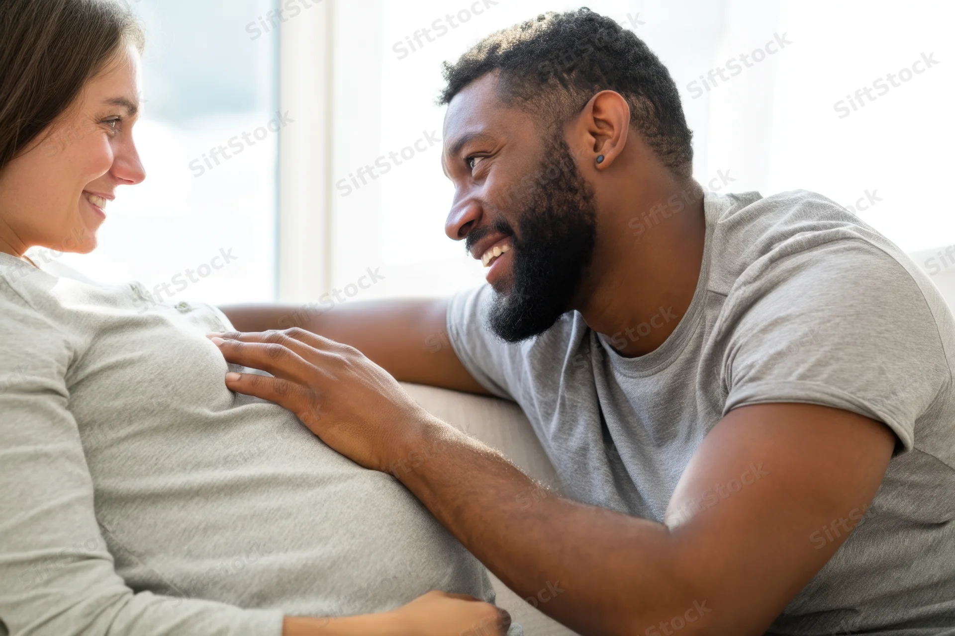 Man smiling warmly at pregnant woman while touching her belly on couch