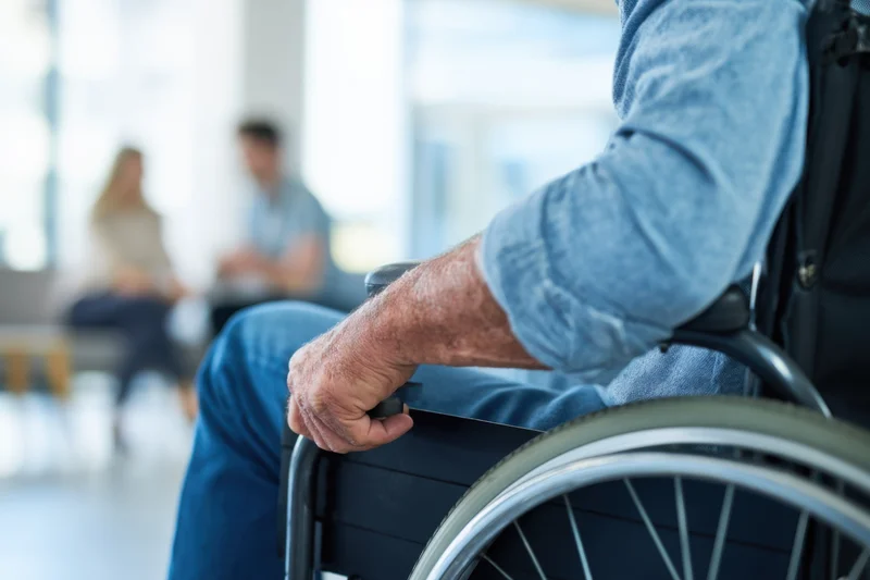 Close-up of a senior man in a wheelchair in a medical waiting room with blurred people in the background