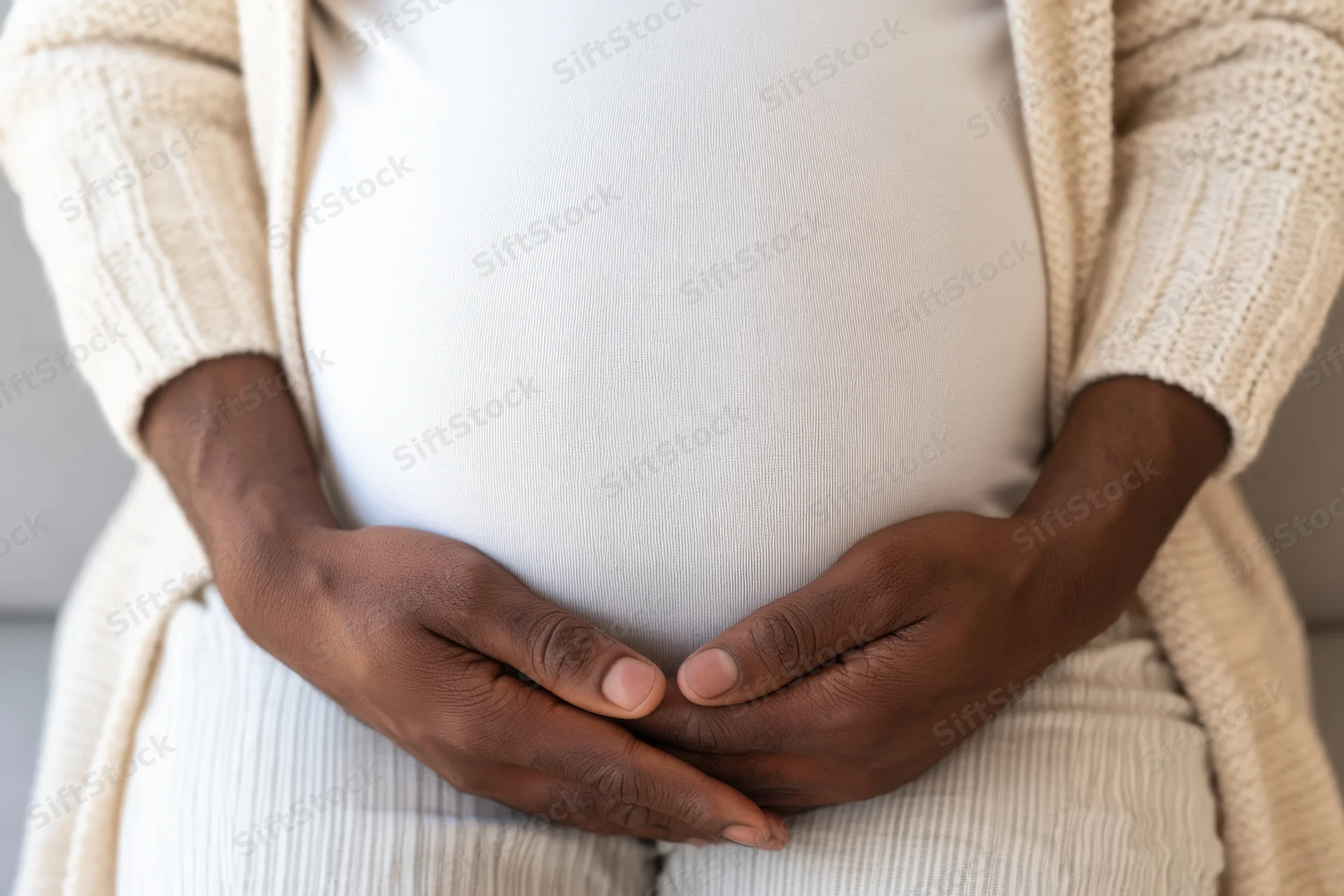 Pregnant woman cradling her belly with hands, wearing ribbed t-shirt and beige cardigan indoors
