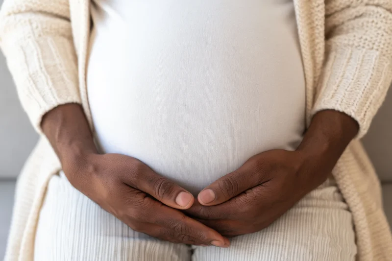 Pregnant woman cradling her belly with hands, wearing ribbed t-shirt and beige cardigan indoors
