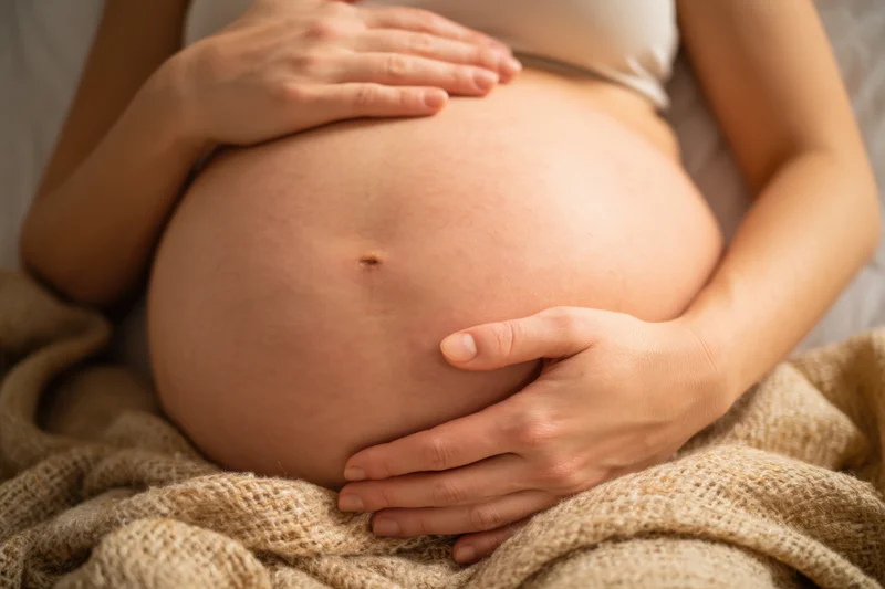 Pregnant woman cradling her belly with hands while lying on bed under knitted blanket