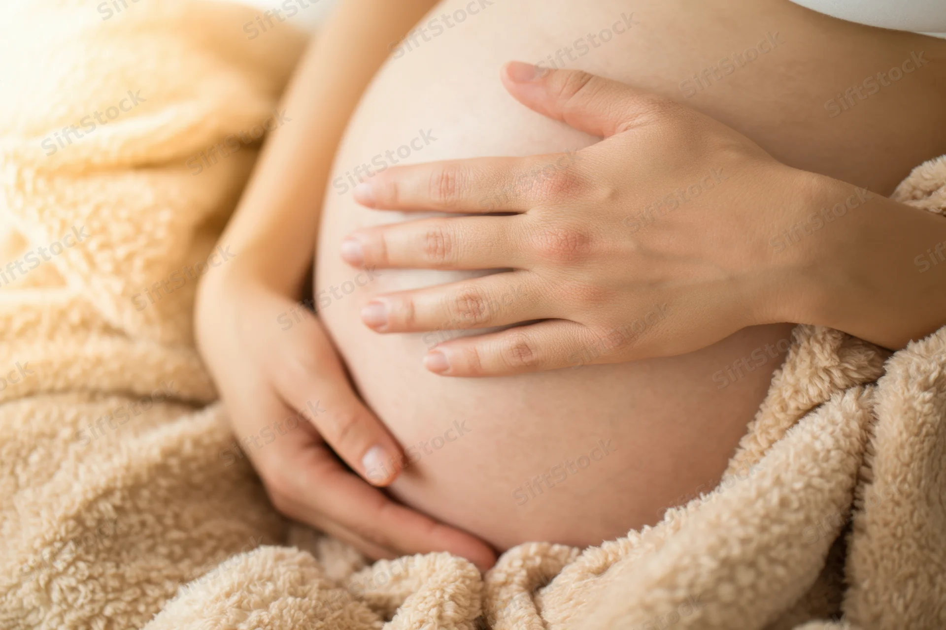 Pregnant woman cradling her belly with hands, wrapped in a soft beige blanket