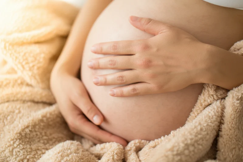 Pregnant woman cradling her belly with hands, wrapped in a soft beige blanket