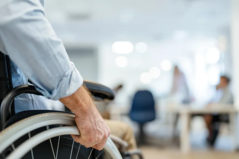 Man in wheelchair gripping wheel in office environment with colleagues at desks in background