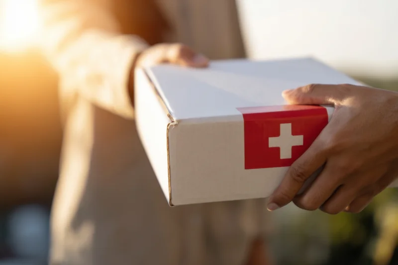 Hands holding a white medical supply box with red cross label outdoors in sunlight