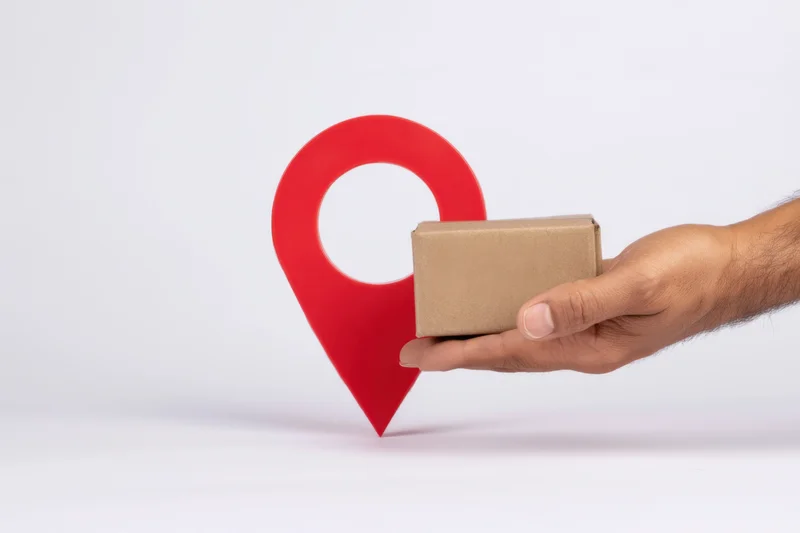 Hand holding small cardboard box in front of red location pin on white background