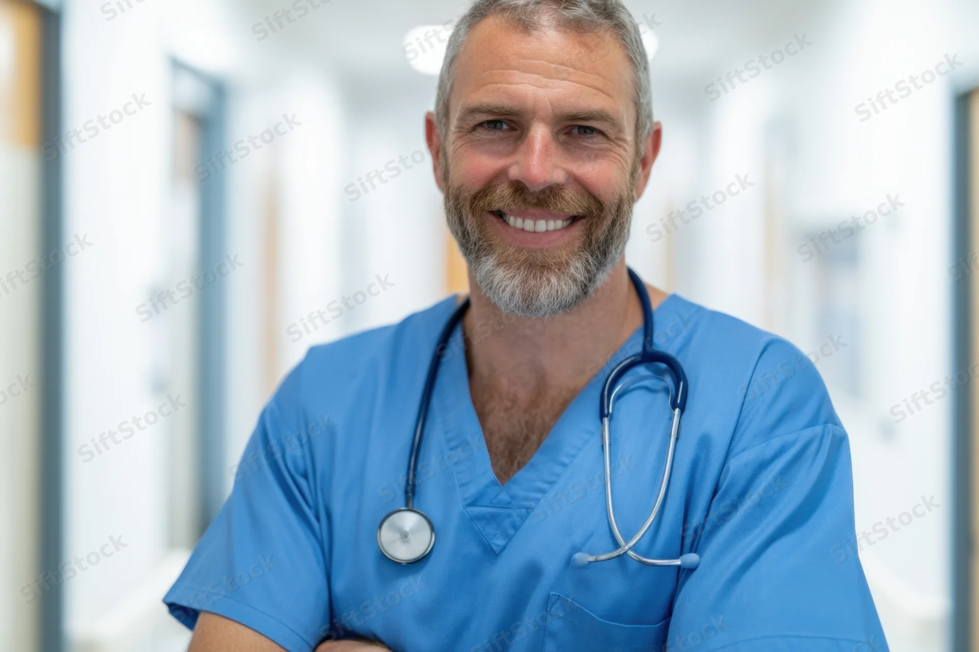 Male doctor in blue scrubs smiling with stethoscope around neck in hospital corridor