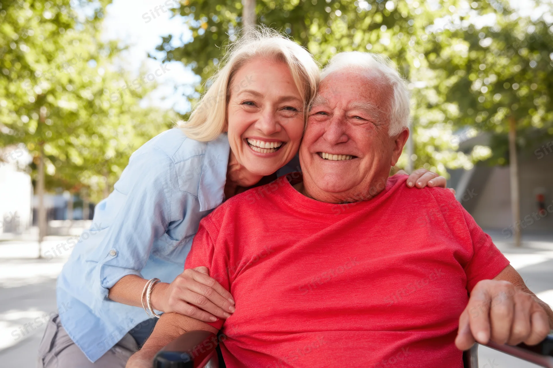 Blonde woman hugging smiling senior man in wheelchair outdoors in park