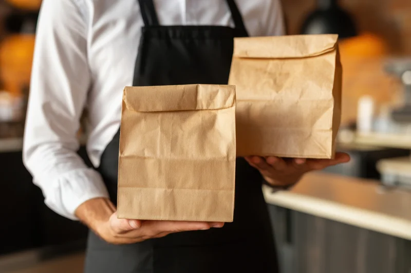 Man in apron holding two paper food bags in a cafe kitchen