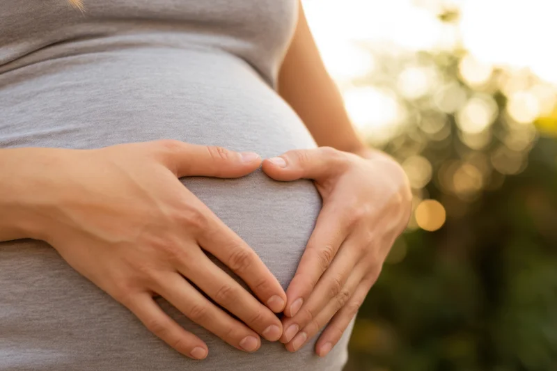 Pregnant woman cradling her belly outdoors with hands forming a heart shape