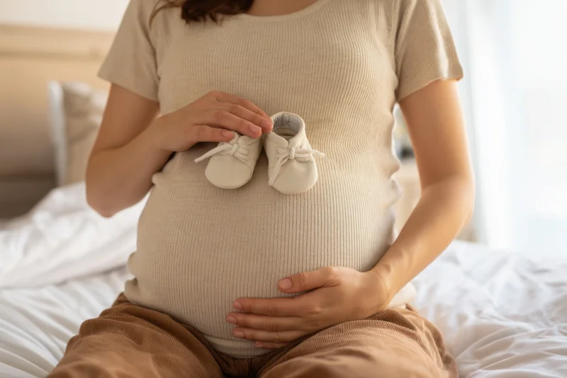 Pregnant woman holding baby shoes on her belly while sitting on bed