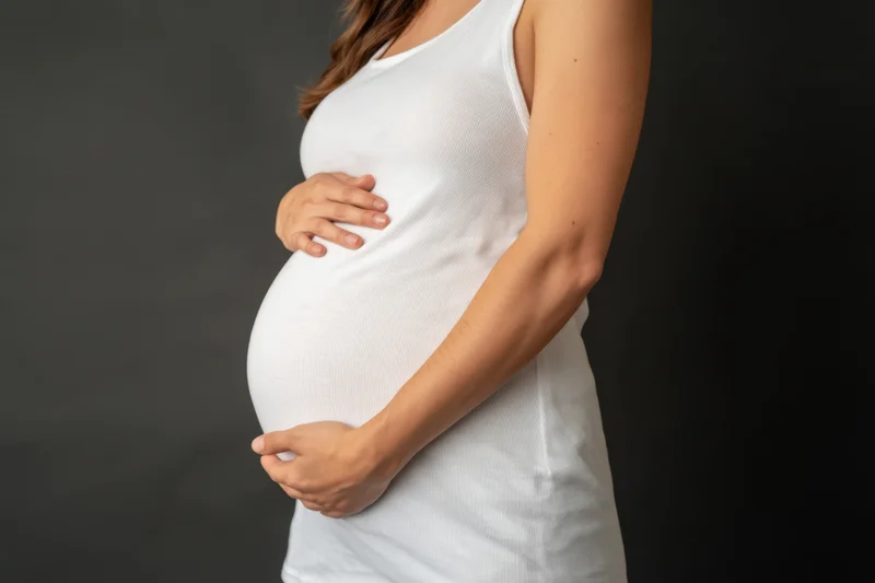 Pregnant woman cradling her belly in a white tank top against a dark studio background