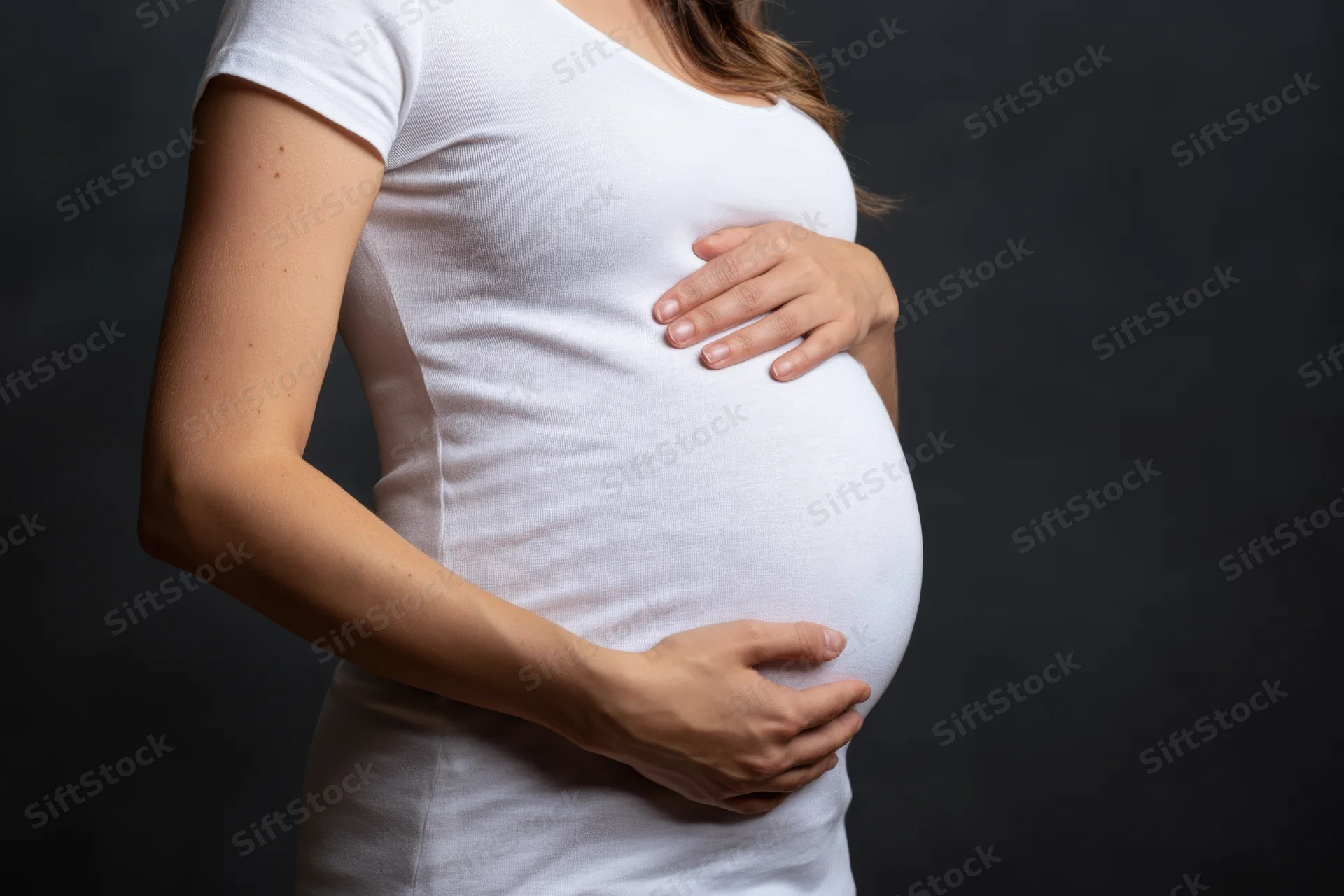 Pregnant woman cradling her belly with both hands against dark background