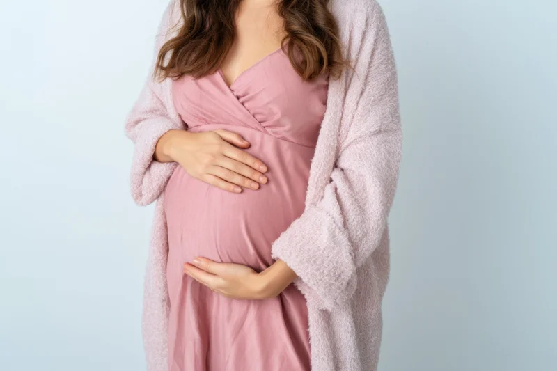 Pregnant woman cradling her belly in a pink dress and robe against a light blue background