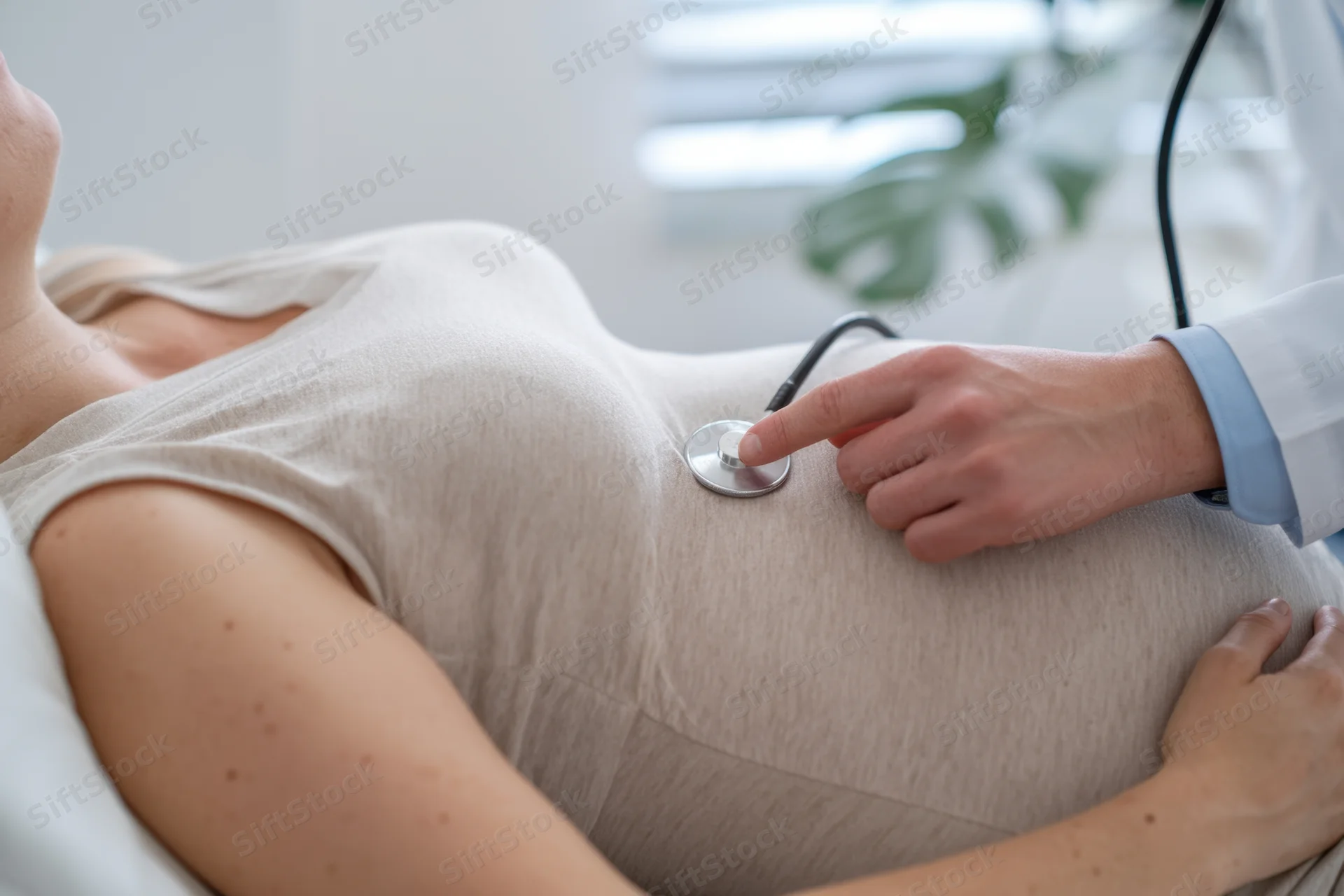 Pregnant woman lying on examination table while doctor uses stethoscope on her abdomen