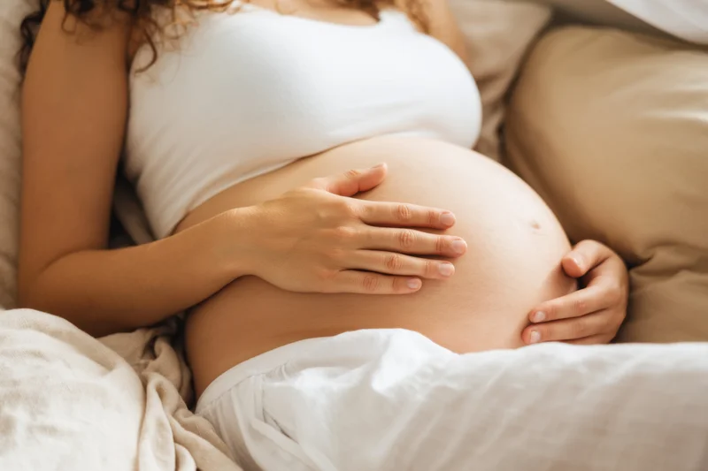 Pregnant woman lying on bed with hands on belly, covered in white sheets