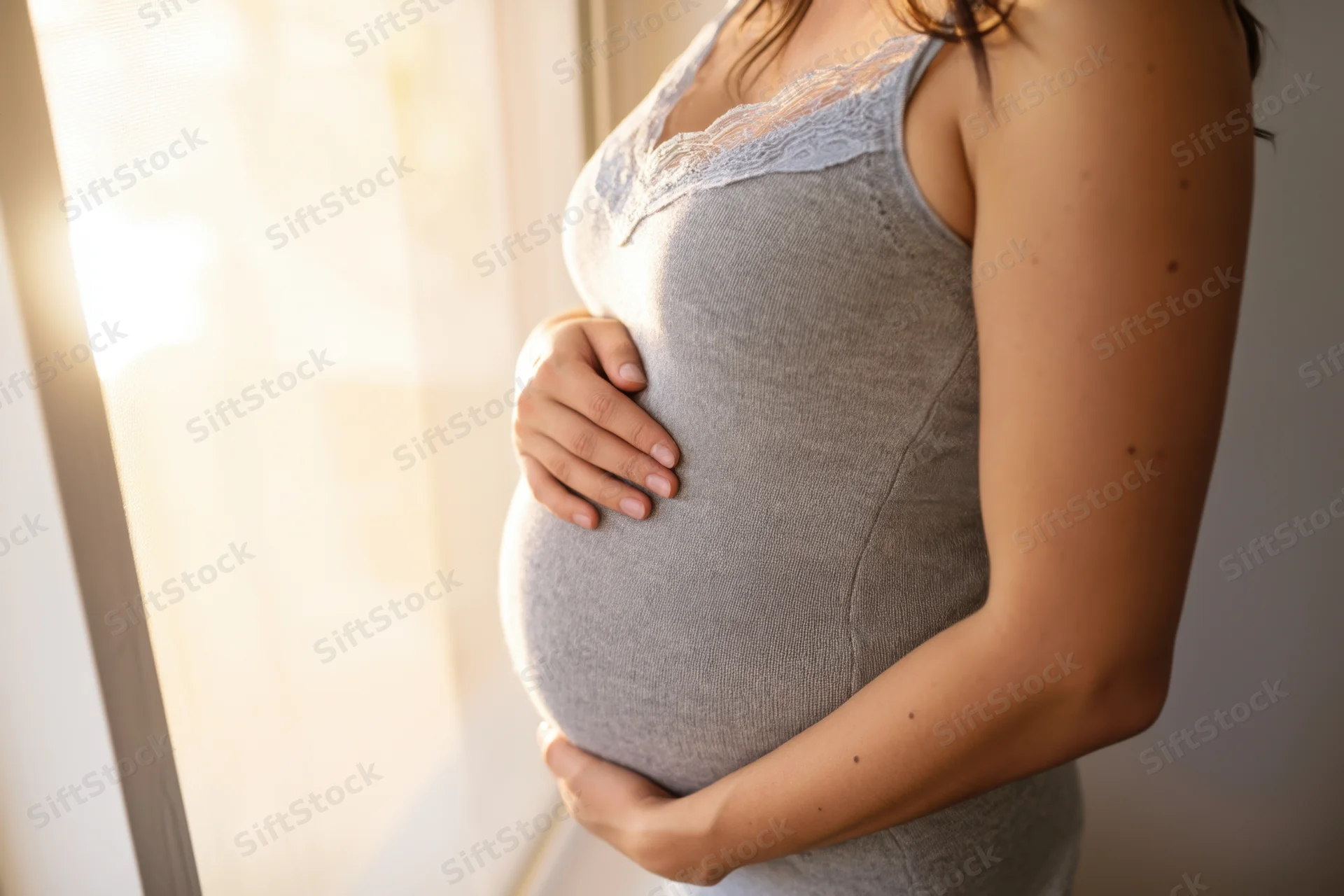 Pregnant woman holding her belly by a sunlit window indoors