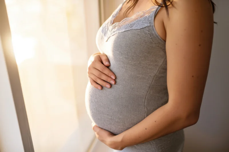 Pregnant woman holding her belly by a sunlit window indoors