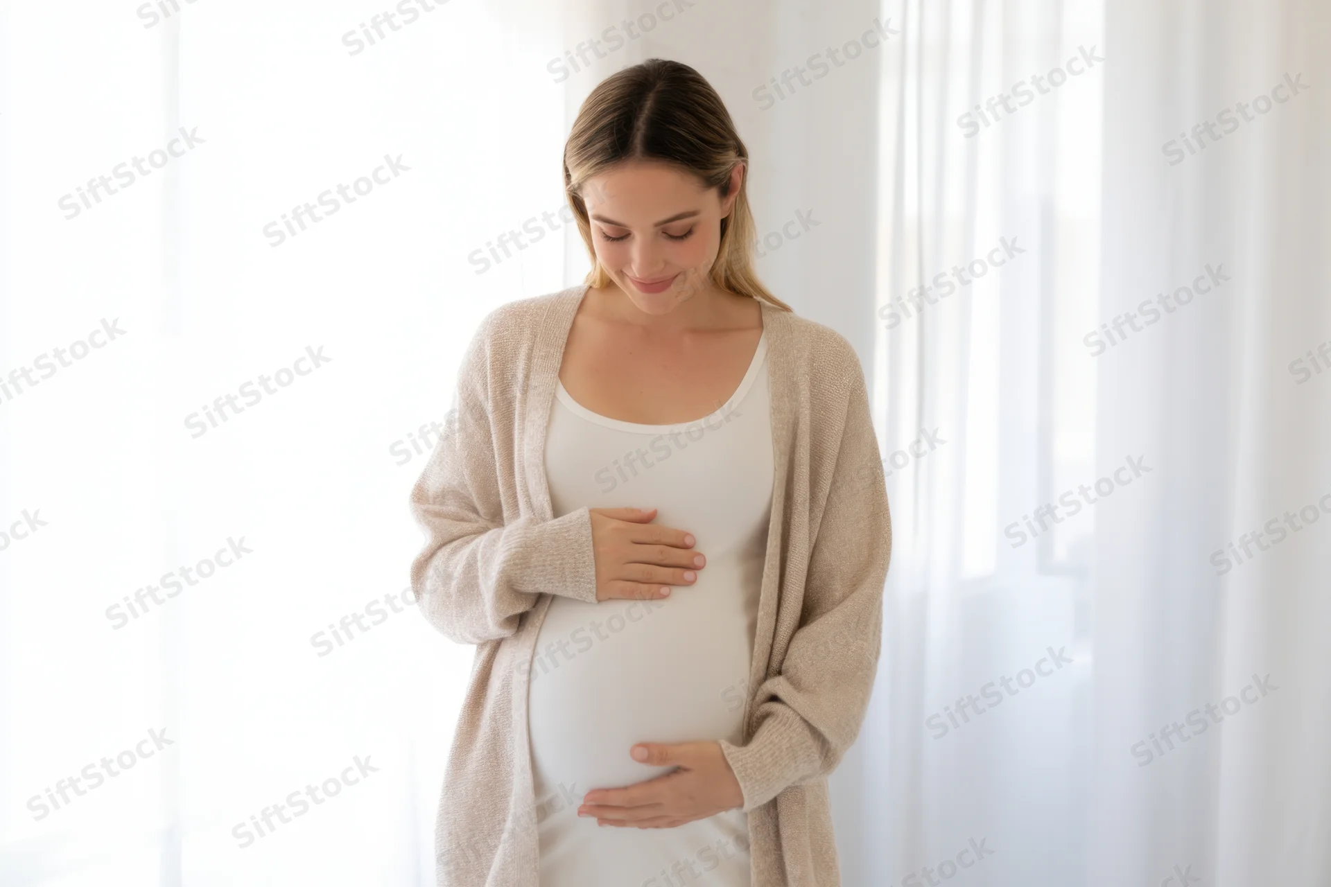 Pregnant woman smiling while touching her belly indoors near a window