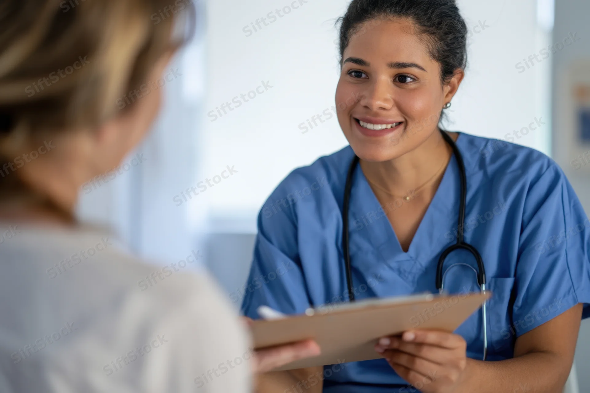 Nurse in blue scrubs smiling while holding clipboard during patient consultation in clinic
