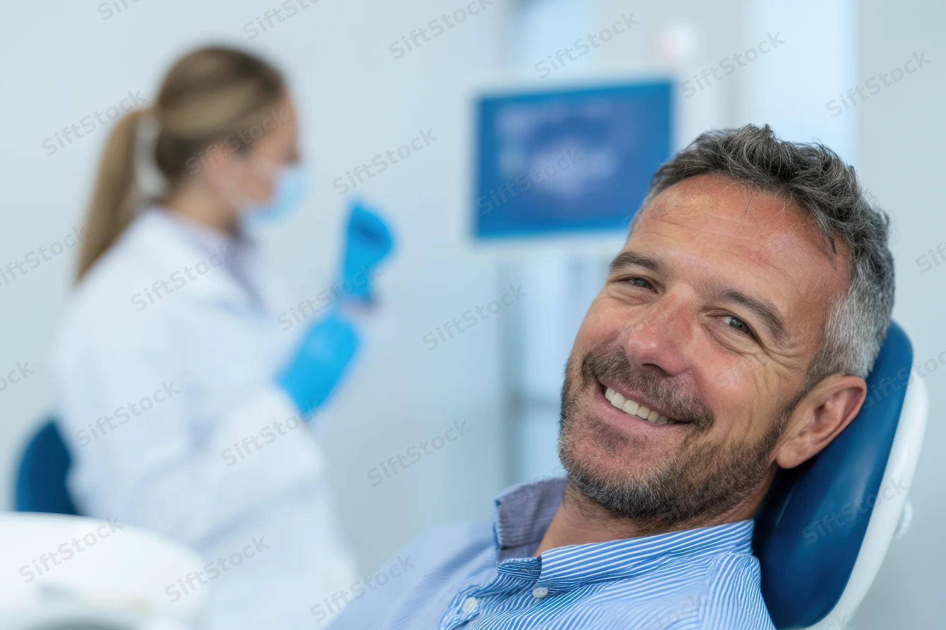 Smiling man in dental chair with dental hygienist in background at clinic