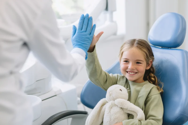 Young girl smiling and high-fiving dentist in dental chair holding stuffed rabbit