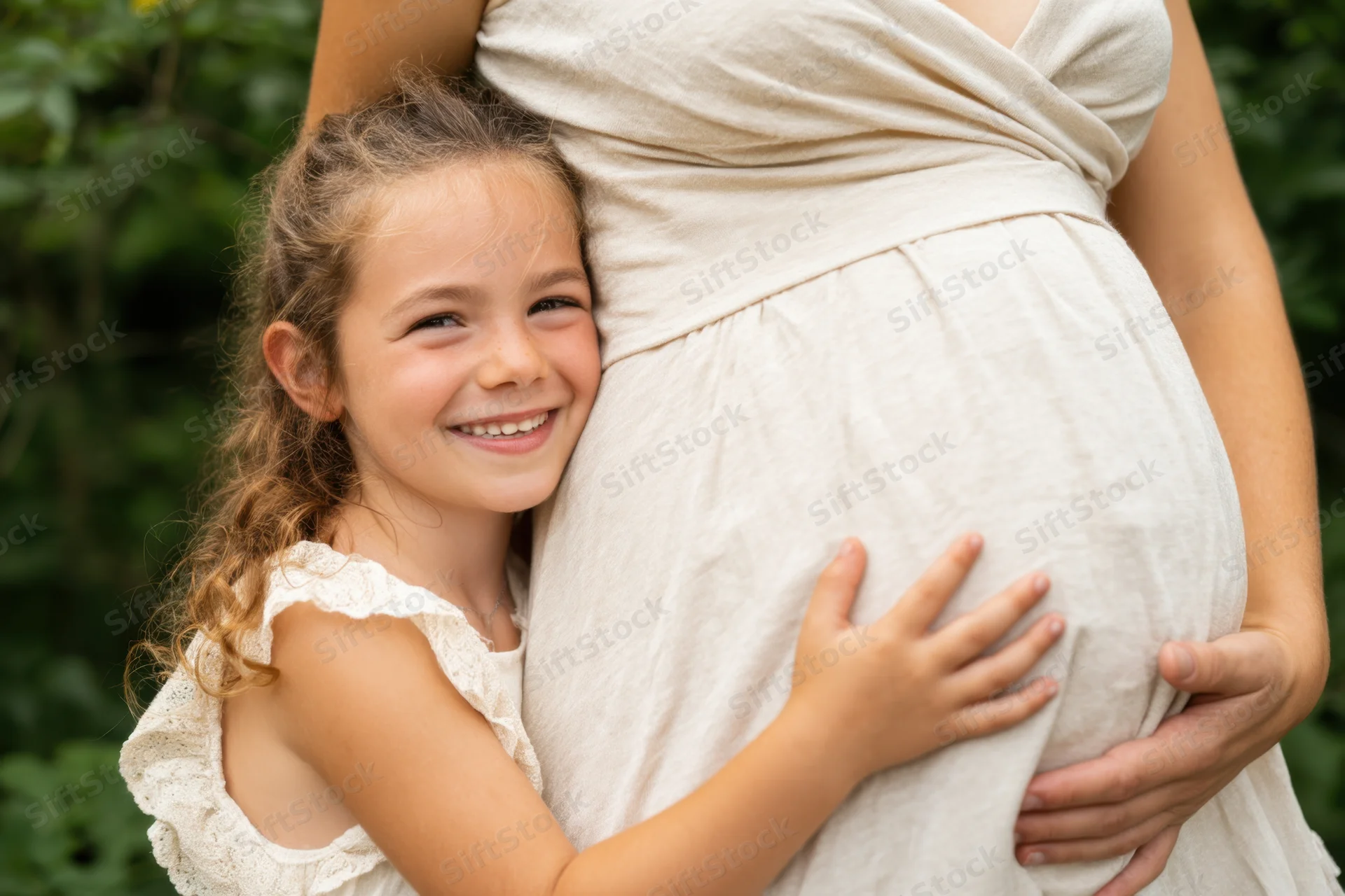 Young girl hugging pregnant woman’s belly in a garden