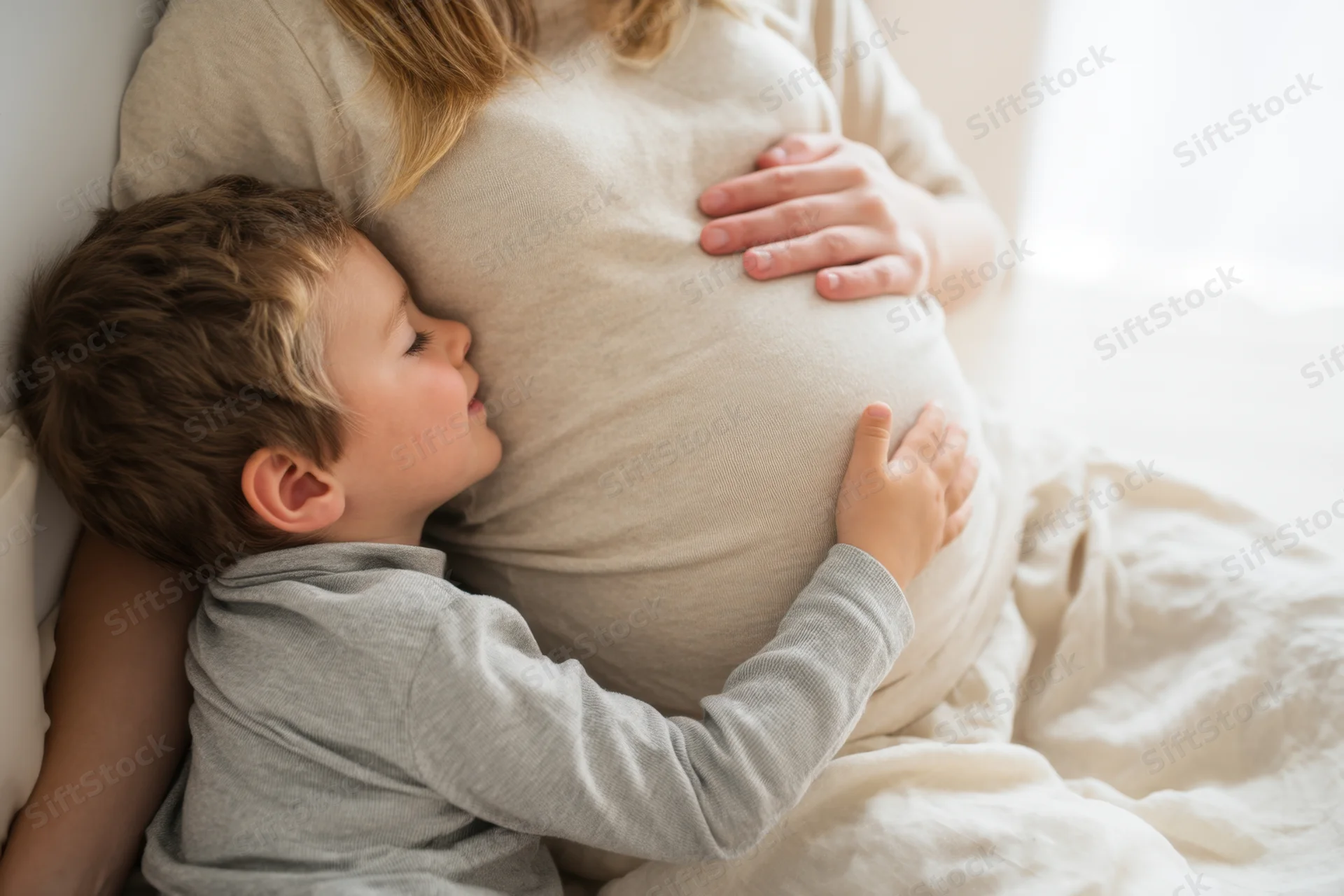 Young boy resting his head on his pregnant mother’s belly while lying in bed together