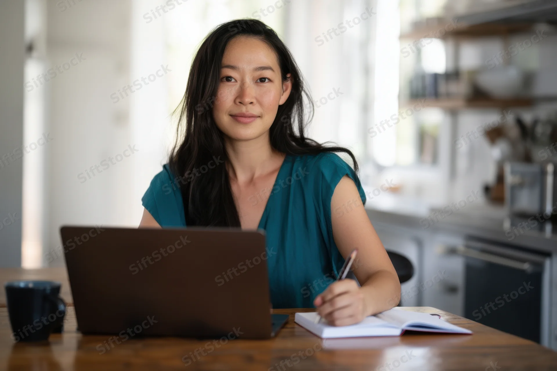 Woman writing in notebook at desk with laptop in kitchen home office