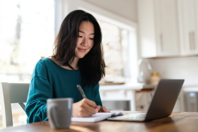 Woman writing in notebook at kitchen table with laptop and coffee mug