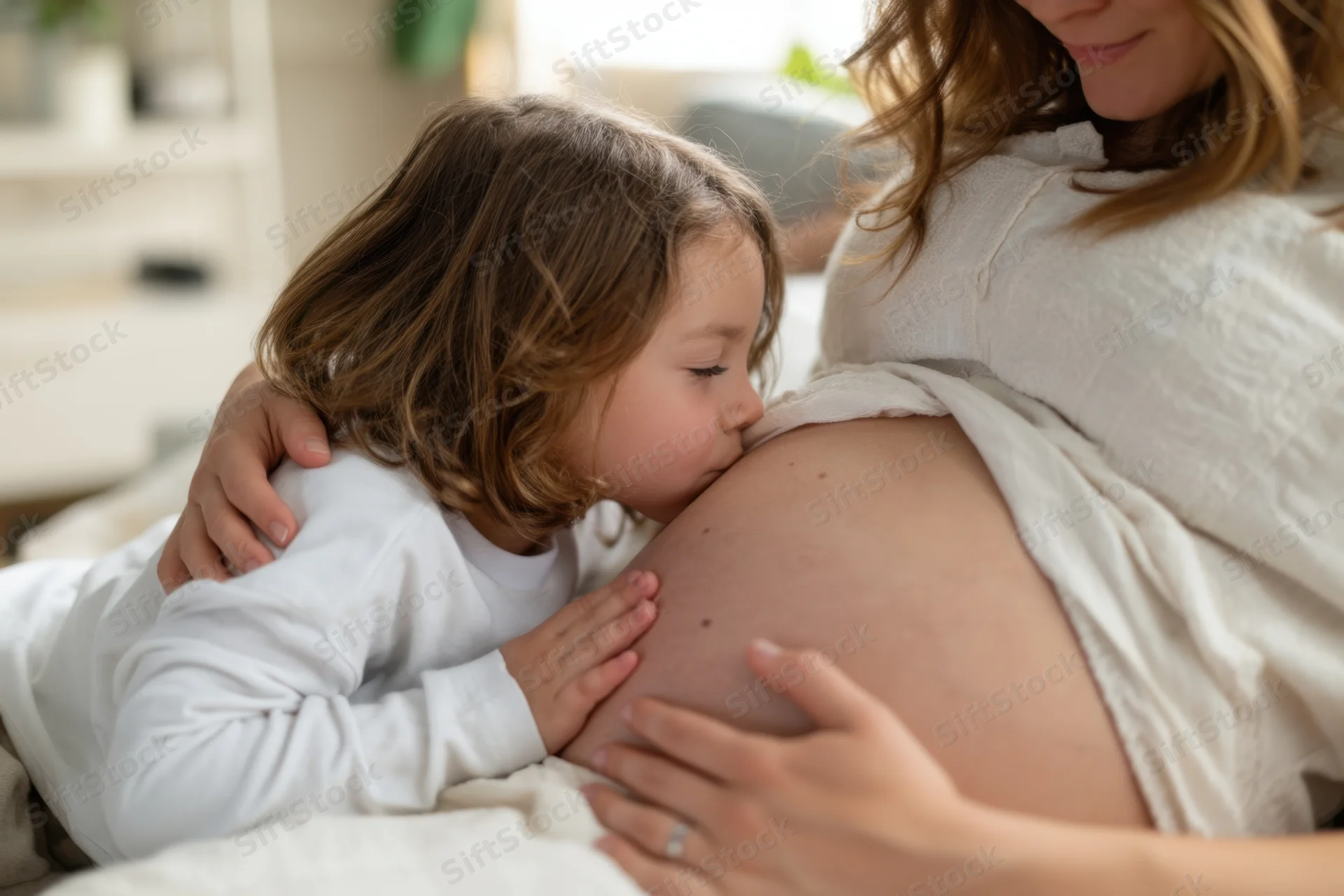 Young girl kissing pregnant woman's belly in a cozy bedroom setting