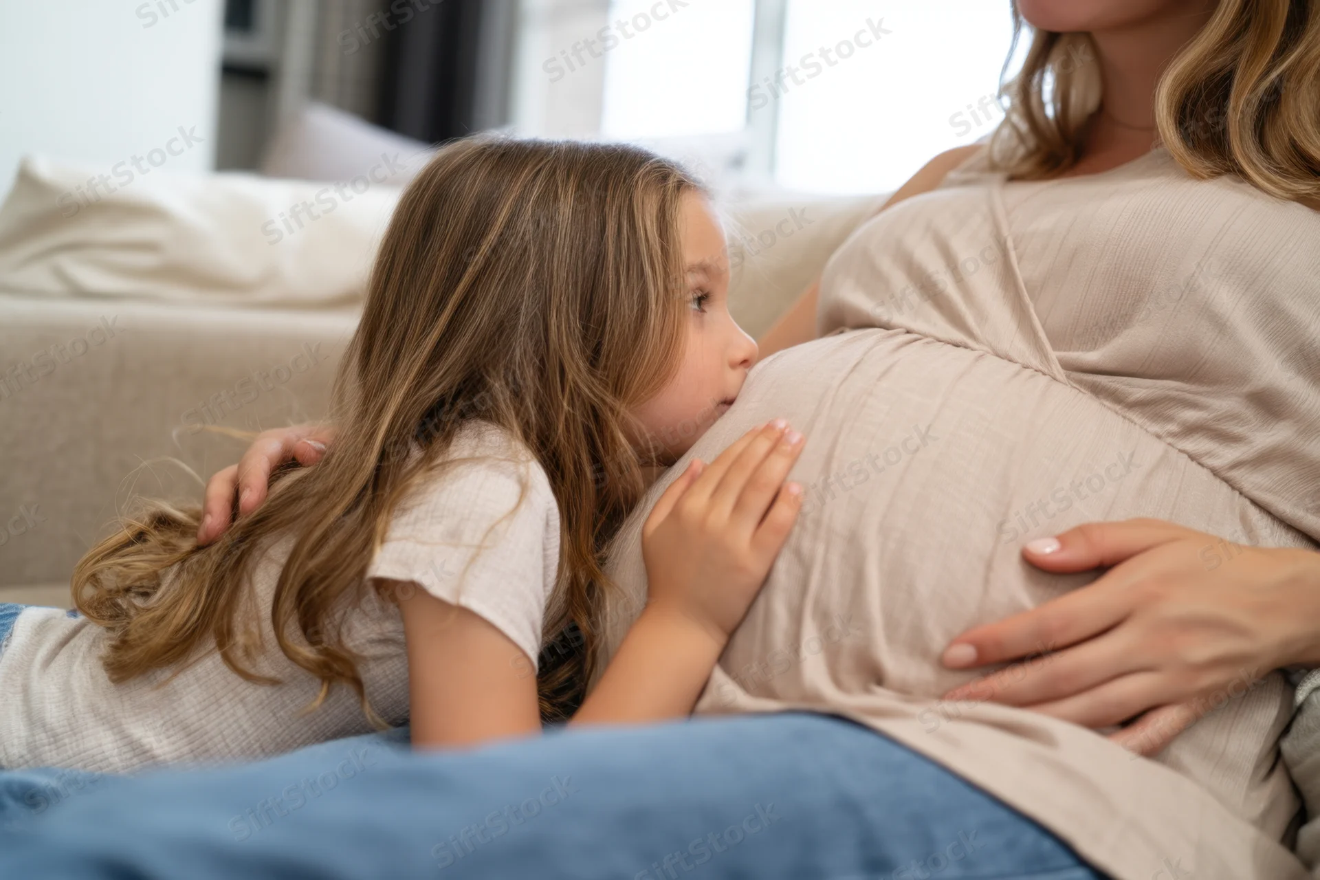 Young girl kissing pregnant woman's belly while lying on couch at home