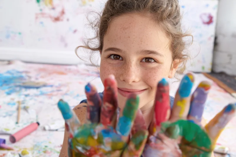 Young girl smiling with paint-covered hands in front of artwork on canvas