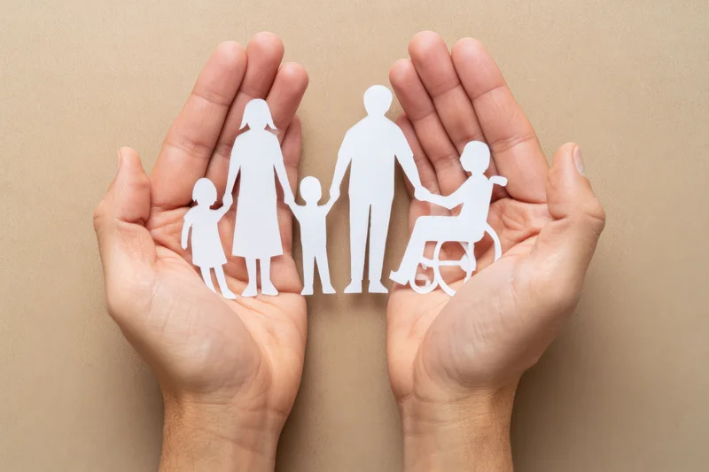 Hands cradling white paper cutouts of a family including a person in a wheelchair against a beige background