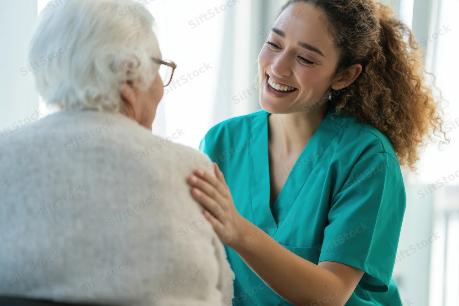 Nurse in teal scrubs smiling and placing hand on senior woman's shoulder during patient interaction in bright indoor setting