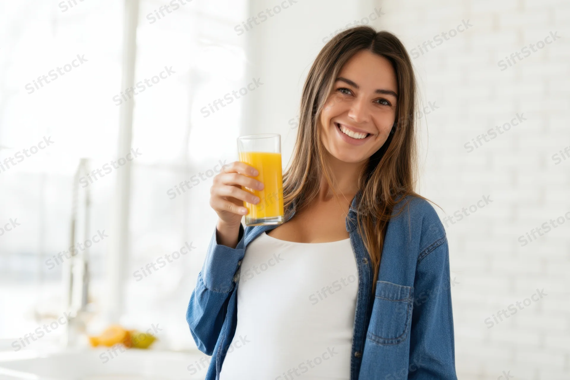Pregnant woman smiling while holding a glass of orange juice in a bright kitchen
