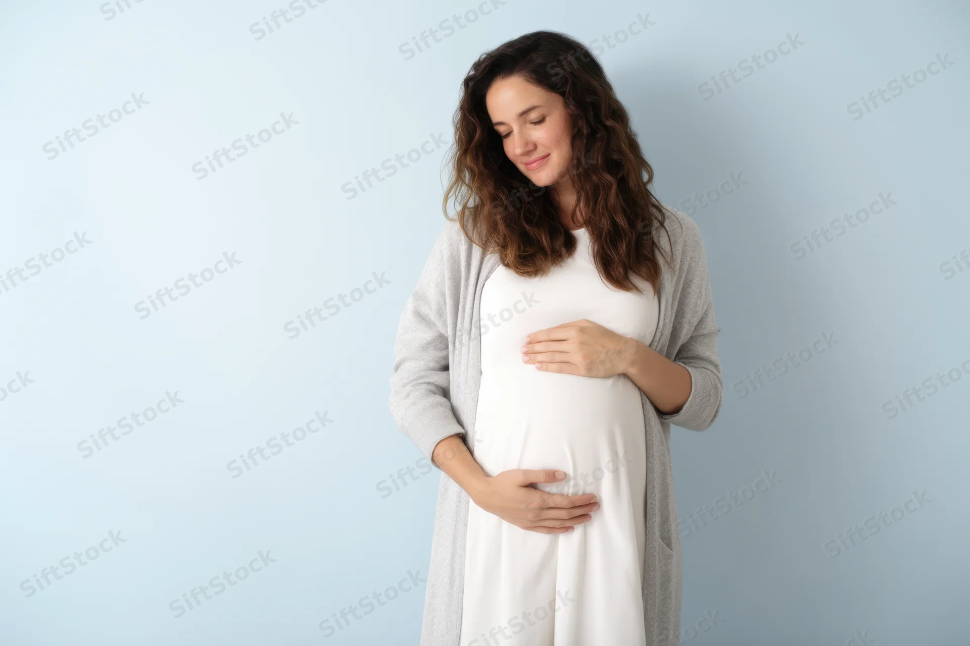 Pregnant woman standing in studio, smiling gently while cradling her belly with both hands, wearing white dress and gray cardigan against light blue background