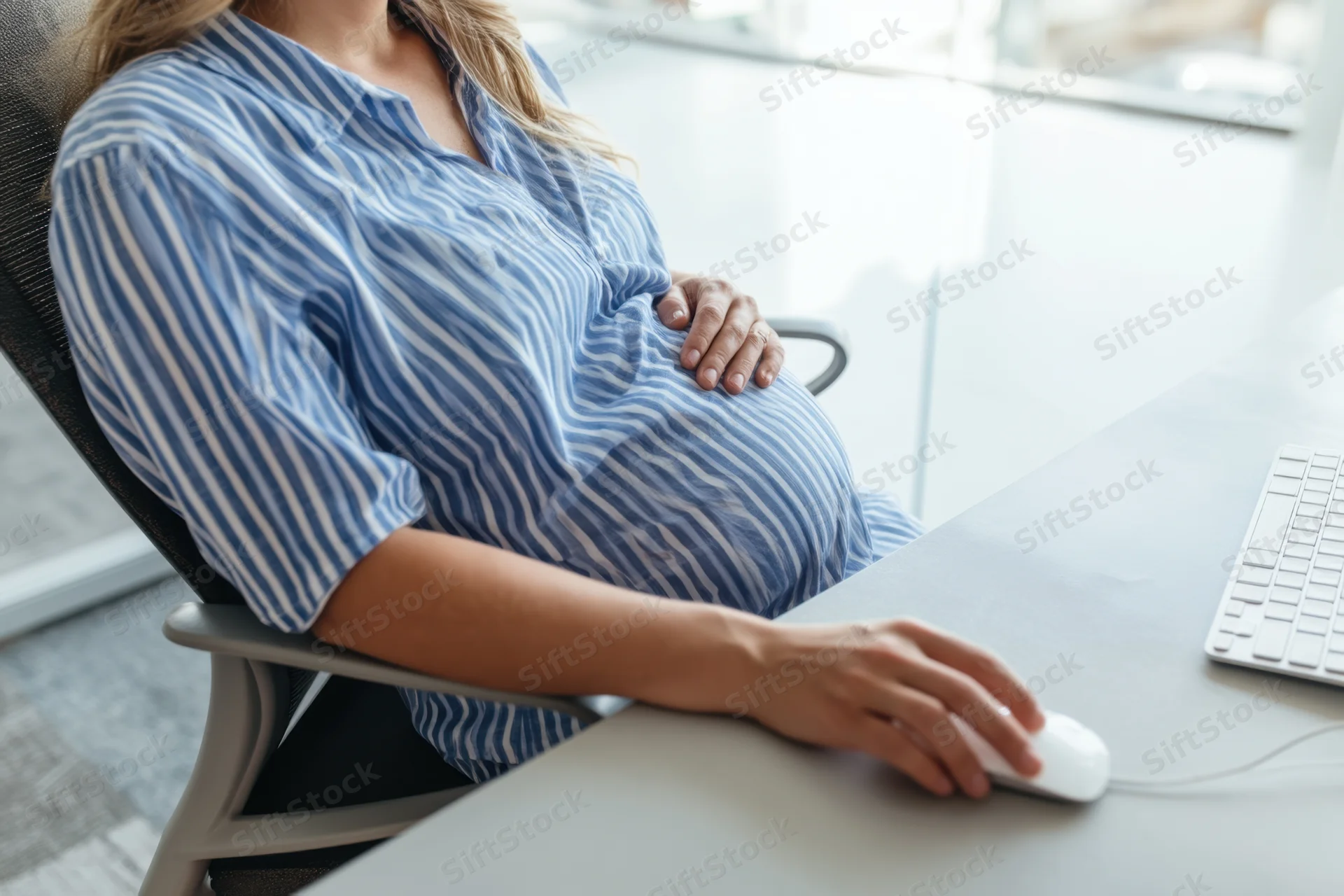 Pregnant woman sitting at office desk using computer mouse while resting hand on belly