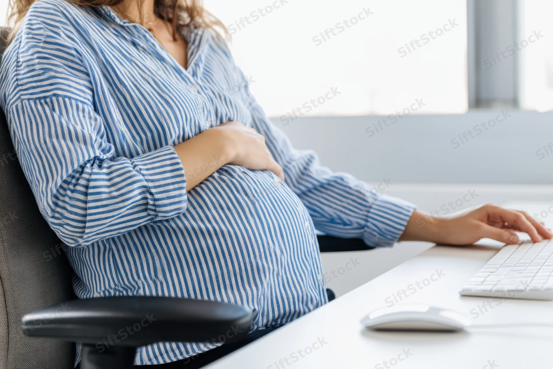 Pregnant woman sitting at office desk using computer while holding her belly