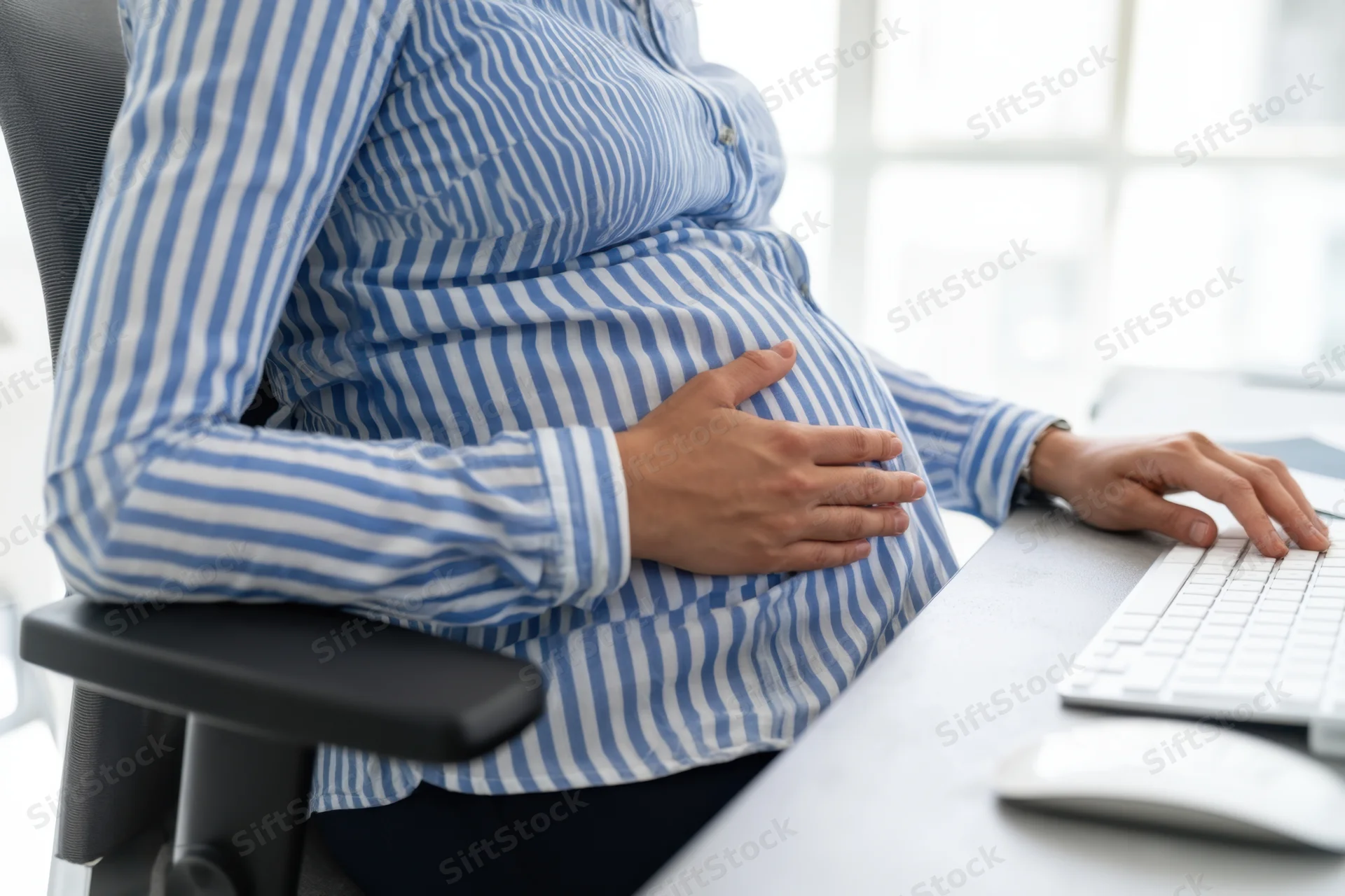 Pregnant woman sitting at office desk typing on keyboard while holding her belly