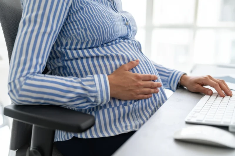 Pregnant woman sitting at office desk typing on keyboard while holding her belly
