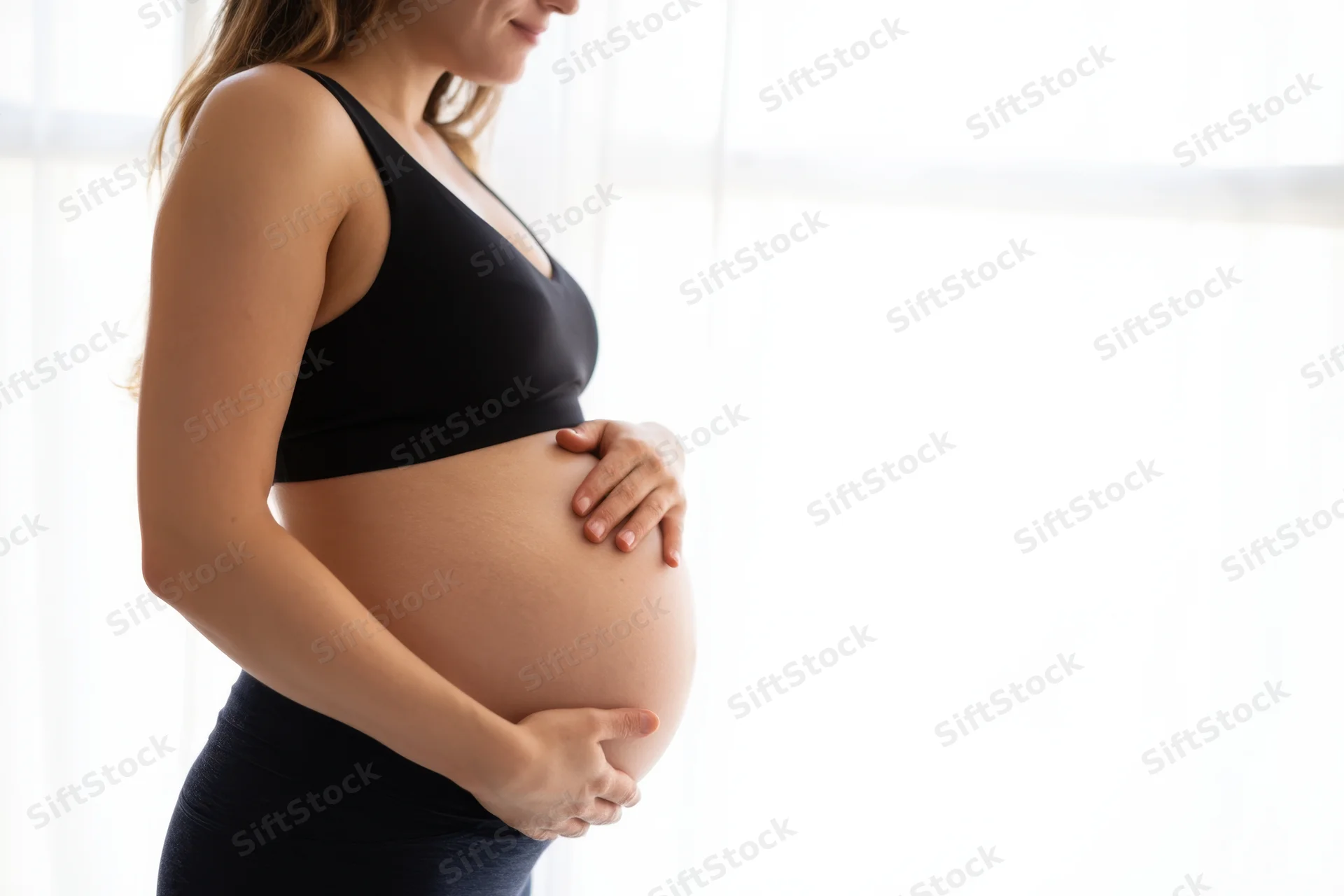 Pregnant woman touching her belly in side profile indoors near a bright window