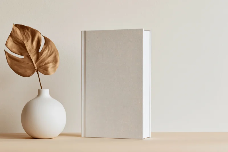 Blank white hardcover book standing upright beside a ceramic vase with a dried monstera leaf on a beige table