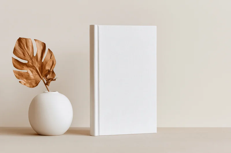 Blank white hardcover book standing upright beside a white vase with a dried monstera leaf on a beige surface