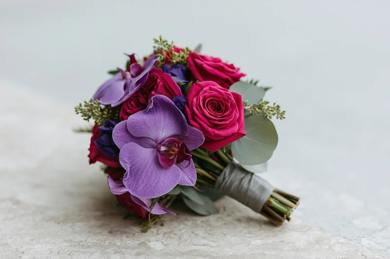 Bridal bouquet of hot pink roses and purple orchids with greenery on a textured surface