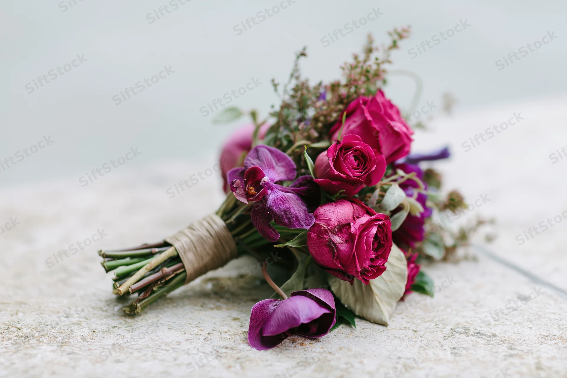 Bridal bouquet of hot pink roses and purple orchids with greenery wrapped in twine resting on stone surface