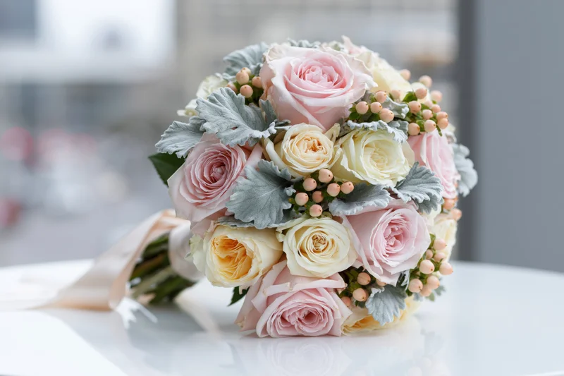 Bridal bouquet of pink and cream roses with dusty greenery resting on white glossy surface